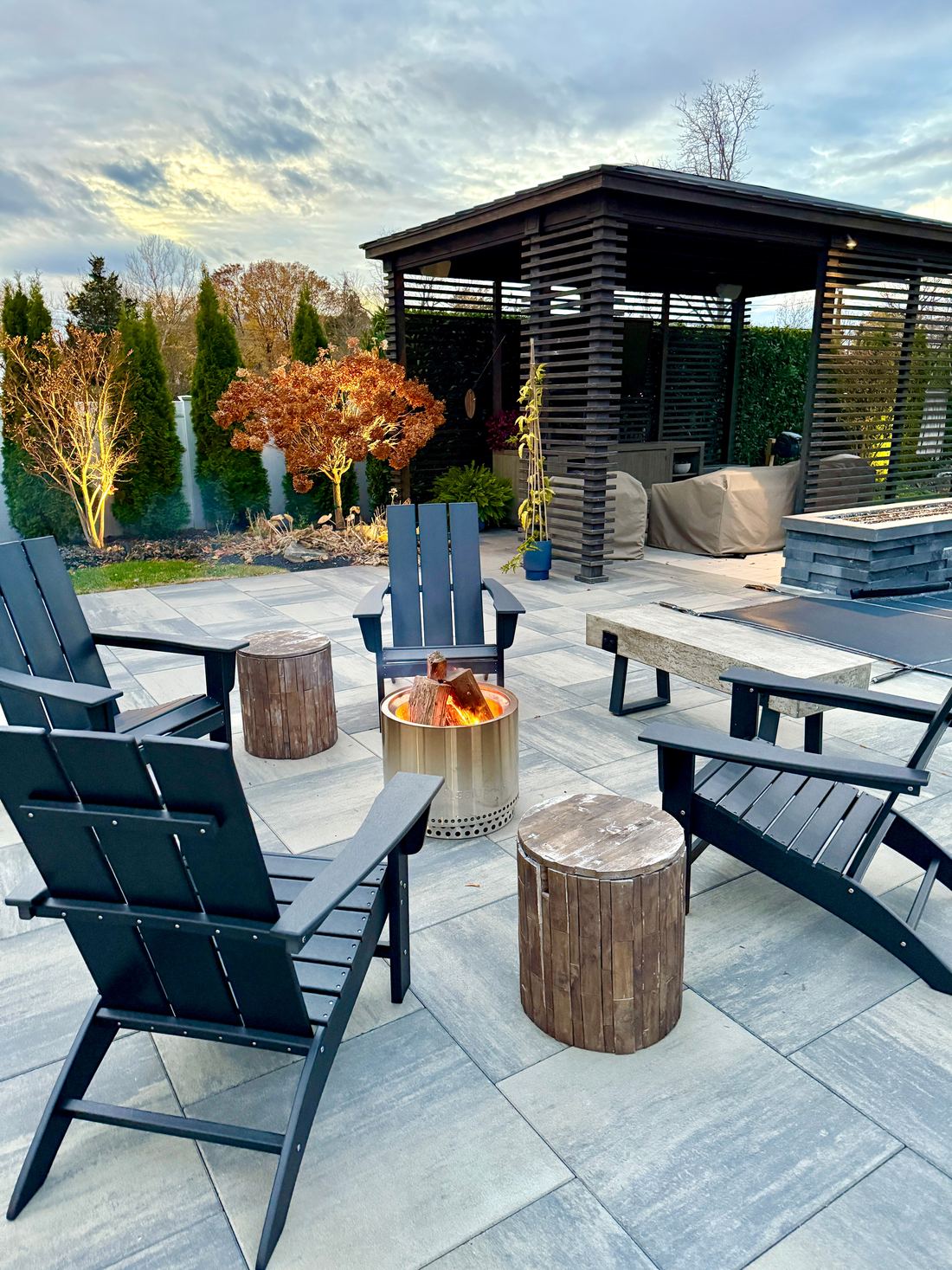 Fire pit with lounge chairs on a stone patio beside a slatted pavilion